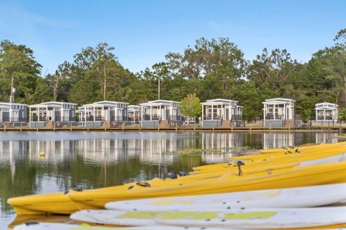 Colorful kayaks and paddleboards in the foreground, with modern cabins and trees reflected in a calm lake.