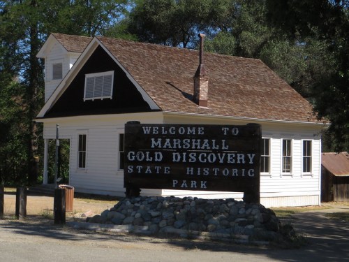 A historic building with a sign reading "Welcome to Marshall Gold Discovery State Historic Park" in a wooded area.