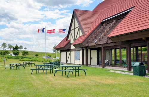 A building with a red roof and flags outside, surrounded by green grass and picnic tables.