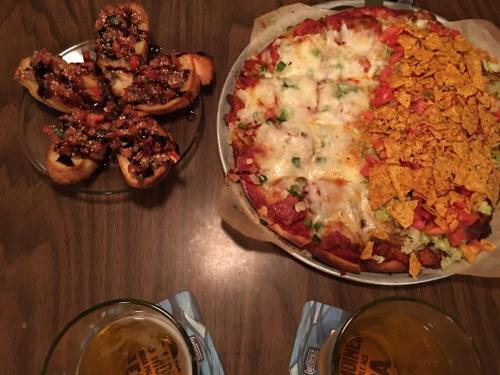 A table with a pizza topped with vegetables and nacho chips, alongside a plate of stuffed bread and two glasses of beer.