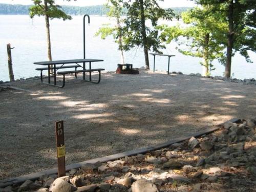 A gravel campsite by a lake, featuring a picnic table, fire pit, and trees in the background.