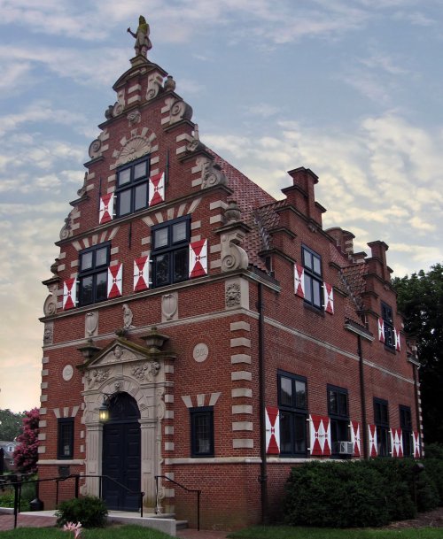 Historic brick building with decorative gables and red and white banners, set against a cloudy sky.