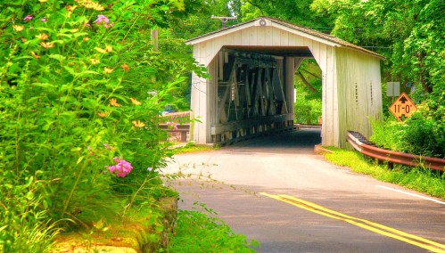 A scenic view of a covered bridge surrounded by lush greenery and colorful flowers, with a winding road leading to it.