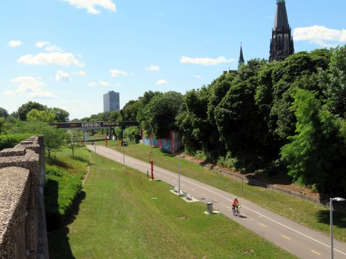 A cyclist rides along a paved path surrounded by greenery, with buildings and a church spire in the background.