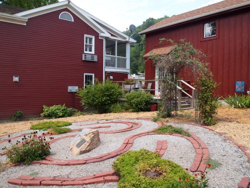 A garden path made of bricks and gravel, surrounded by greenery, with two red buildings in the background.