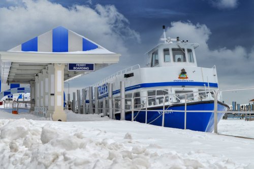 A blue and white ferry docked at a snowy terminal under a cloudy sky, with a sign for ferry boarding visible.