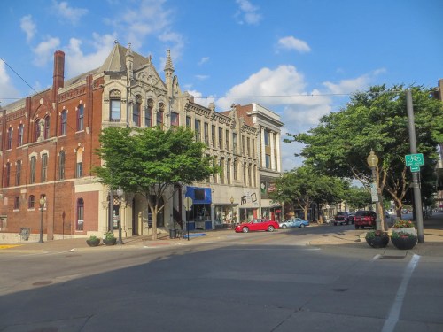 Historic buildings line a street under a blue sky, with trees and a bike lane visible in the foreground.