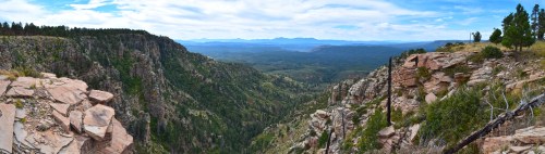 A panoramic view of a lush valley surrounded by rocky cliffs and distant mountains under a blue sky.