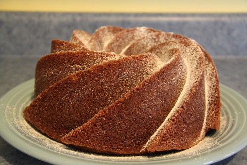 A beautifully baked chocolate bundt cake with a textured swirl, dusted lightly with flour, on a blue plate.