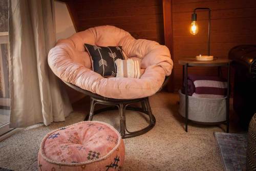 Cozy corner featuring a large pink chair, a side table with a lamp, and a decorative pouf on a carpeted floor.