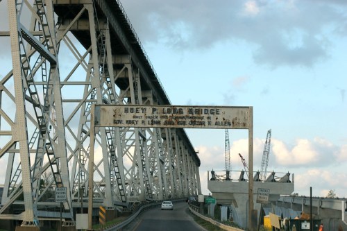 A view of the Huey P. Long Bridge with a vehicle driving underneath and construction cranes in the background.