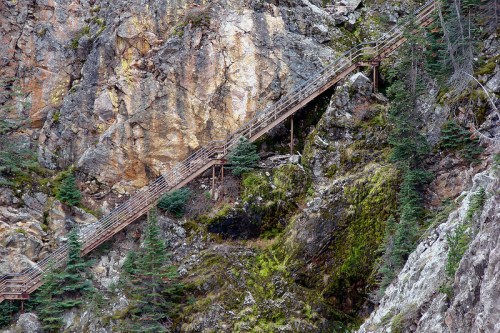 A wooden staircase winds up a rocky mountainside, surrounded by greenery and rugged terrain.