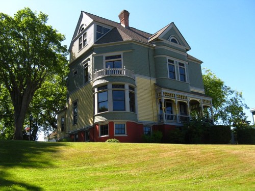 A large, historic house with green siding and a red base, surrounded by lush grass and trees under a clear blue sky.