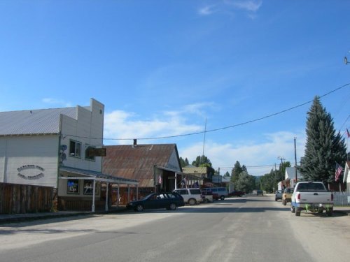 A quiet small-town street with rustic buildings, parked cars, and a clear blue sky.