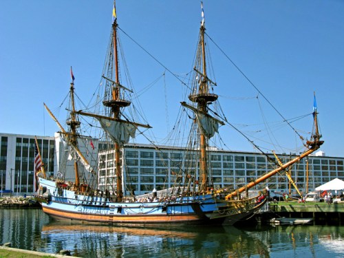 A historic tall ship with three masts docked in a marina, surrounded by modern buildings and clear blue skies.