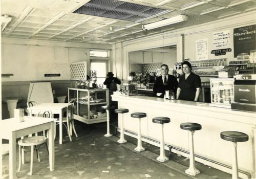 A vintage diner interior with a counter, stools, and two staff members standing behind the bar.