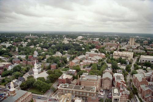 Aerial view of a cityscape with green trees, buildings, and cloudy skies in the background.