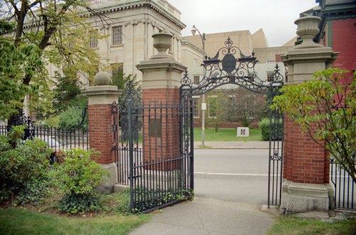 A decorative wrought iron gate opens to a brick pathway lined with greenery, leading to a building in the background.