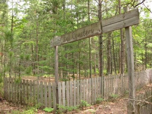A wooden sign reading "Bell Cemetery" stands at the entrance to a wooded area surrounded by a picket fence.