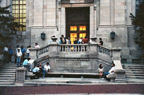 A group of people gathers on the steps of a grand building, with warm lights glowing from the entrance.