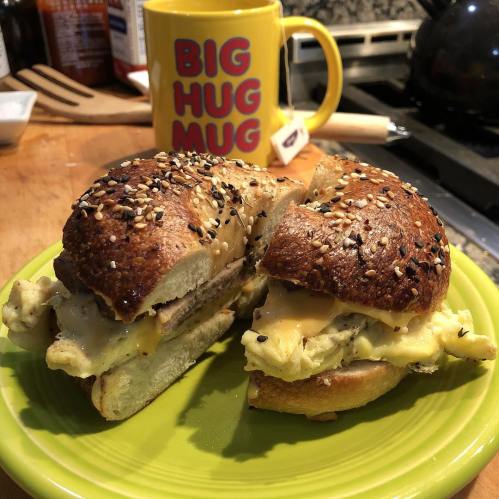 A toasted bagel sandwich filled with scrambled eggs and cheese, served on a green plate with a colorful mug in the background.