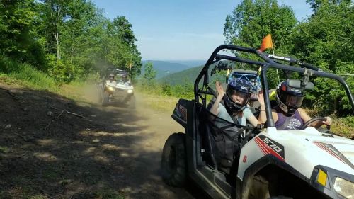 Two off-road vehicles drive on a dirt trail through a forested area, with one passenger waving happily.