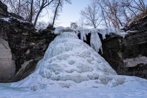 A frozen waterfall cascading down rocky cliffs, surrounded by snow and bare trees under a clear blue sky.