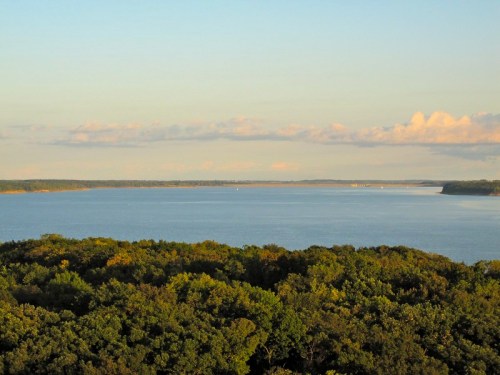 A serene view of a calm lake surrounded by lush greenery under a clear sky with soft clouds.