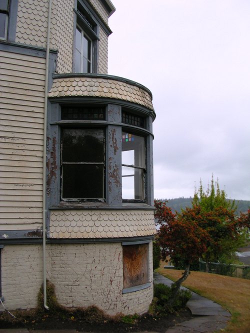 A weathered house corner with a curved window, peeling paint, and a small tree nearby under a cloudy sky.