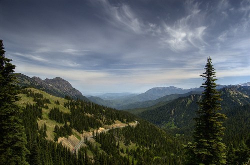 A scenic mountain landscape with lush green forests, winding roads, and a dramatic sky filled with clouds.