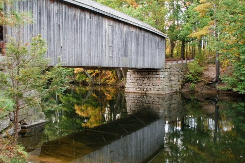 A wooden covered bridge spans a calm river, reflecting in the water, surrounded by autumn foliage.