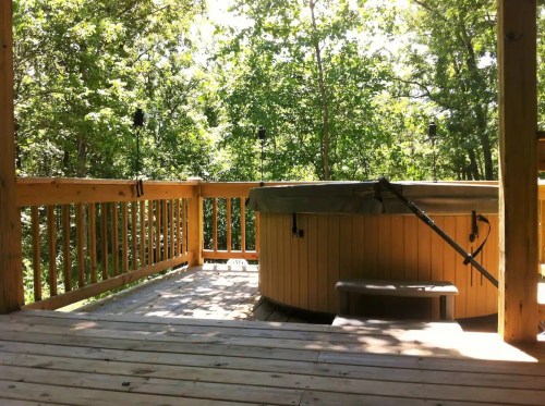A wooden deck with a hot tub surrounded by lush green trees on a sunny day.