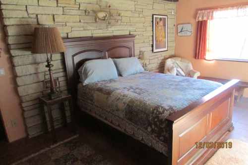 Cozy bedroom featuring a wooden bed, stone wall, and a chair by the window with warm lighting.