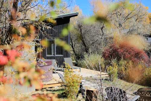 A cozy cabin surrounded by colorful autumn foliage and greenery under a clear blue sky.