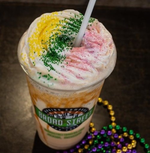 A colorful milkshake topped with whipped cream and sprinkles, served in a cup from Broad Street. Mardi Gras beads nearby.