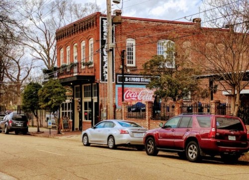 Historic brick building with a Coca-Cola sign, surrounded by trees and parked cars on a quiet street.