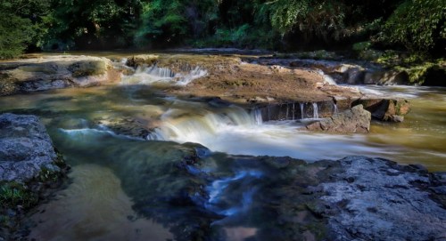 A serene river flows over rocky terrain, with gentle waterfalls surrounded by lush greenery.