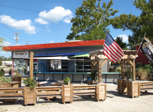 A colorful storefront with an orange awning, American flags, and wooden seating in front, under a blue sky.