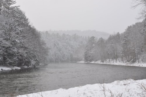 A snowy river scene surrounded by trees, with a misty landscape in the background.