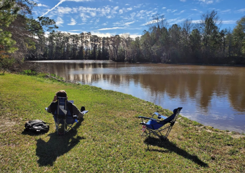 Two camping chairs by a calm river, with a person sitting in one chair, surrounded by trees and a clear blue sky.