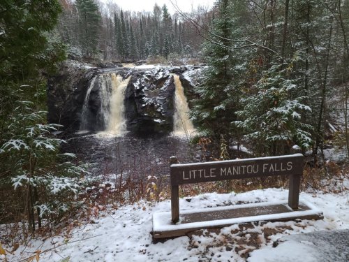A snowy scene of Little Manitou Falls, with two waterfalls cascading into a dark pool surrounded by evergreen trees.