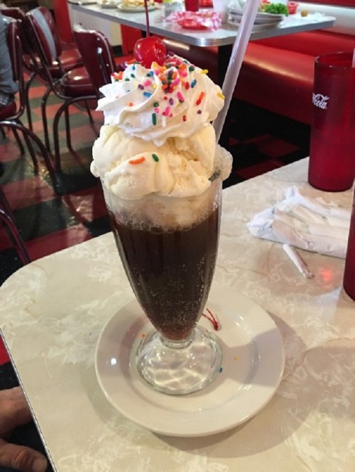 A tall glass of root beer float topped with whipped cream, a cherry, and colorful sprinkles on a retro diner table.