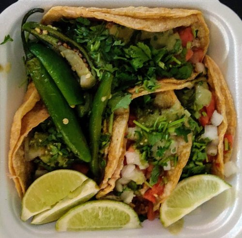 Three tacos filled with fresh toppings, served with green chilies and lime wedges in a white container.