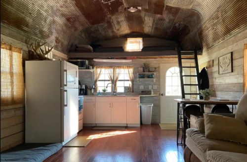 Cozy interior of a small cabin featuring a kitchen, dining area, and a lofted sleeping space with rustic decor.