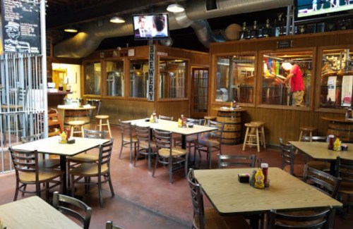 Interior of a brewery with wooden tables, chairs, and a bar area; brewing equipment visible in the background.