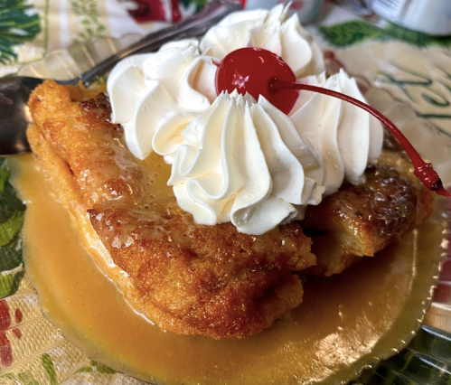 A slice of dessert topped with whipped cream and a cherry, sitting in a sweet sauce on a decorative plate.