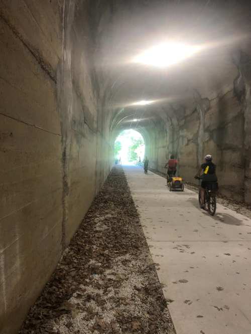 Two cyclists ride through a tunnel with a light at the end, surrounded by leaves on the ground.