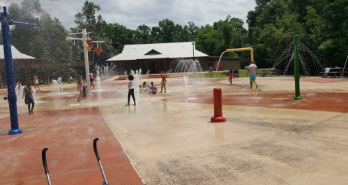 Children playing in a splash pad with water features on a sunny day, surrounded by trees and a building in the background.