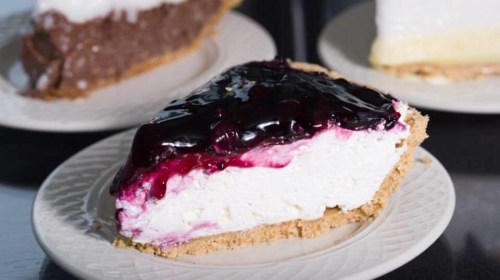 A slice of cheesecake topped with blueberry sauce on a decorative plate, with other pie slices in the background.