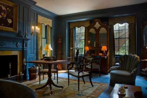 A cozy, elegantly furnished room with dark blue walls, a fireplace, and vintage furniture, illuminated by soft lighting.